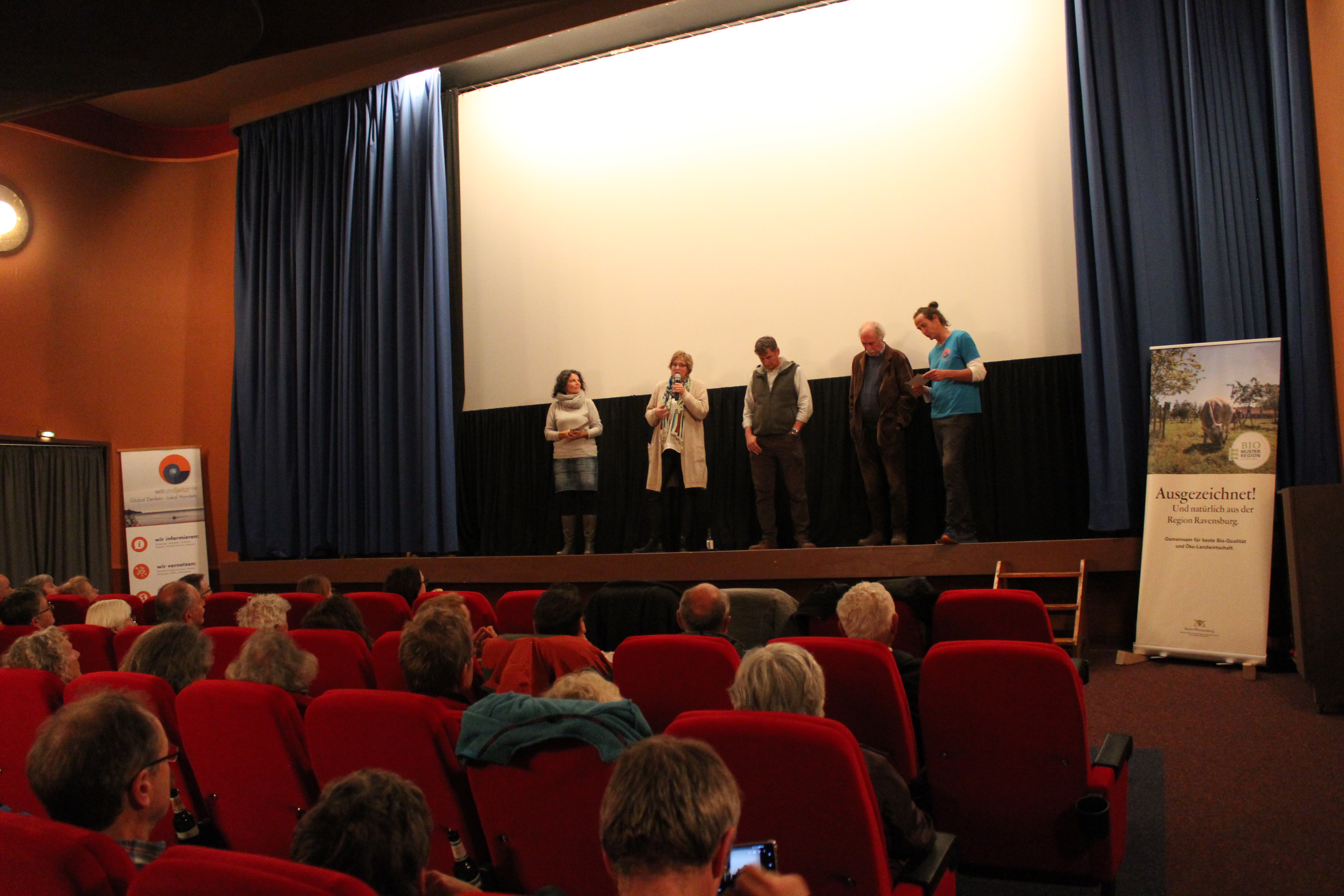 Man sieht die drei Gesprächsteilnehmer mit den beiden Moderatoren auf der Bühne im Kino in Leutkirch. Davor der beinahe vollbesetzte Kinosaal. 
